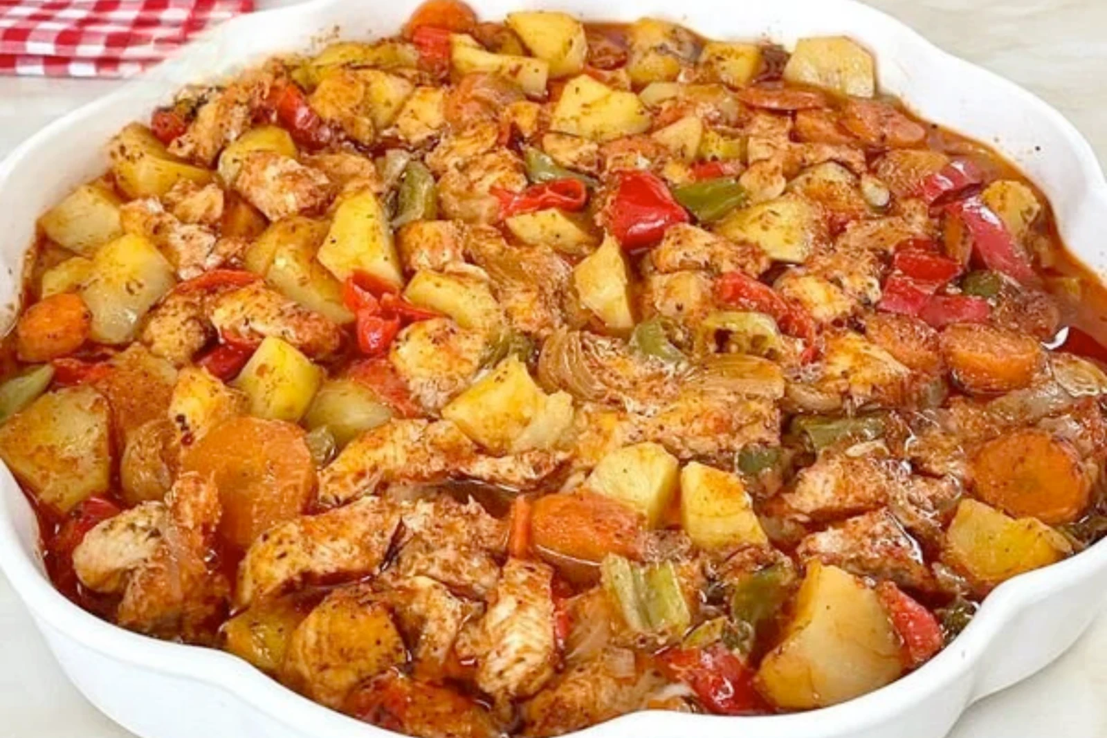 A close-up of a baked Turkish chicken and vegetable casserole in a white dish, showing golden-brown potatoes, carrots, red and green peppers, and seasoned chicken in a flavorful sauce.